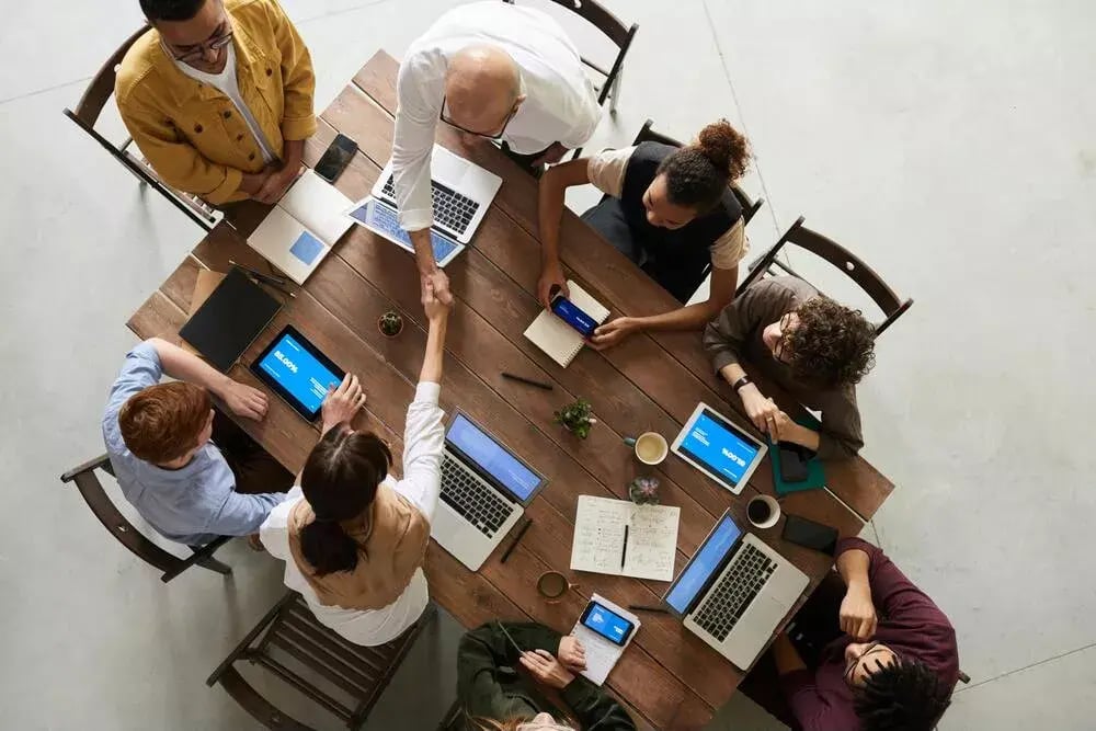 A group of five people is sitting around a wooden table, each with a laptop in front of them; they appear to be in a meeting or a collaborative work session.