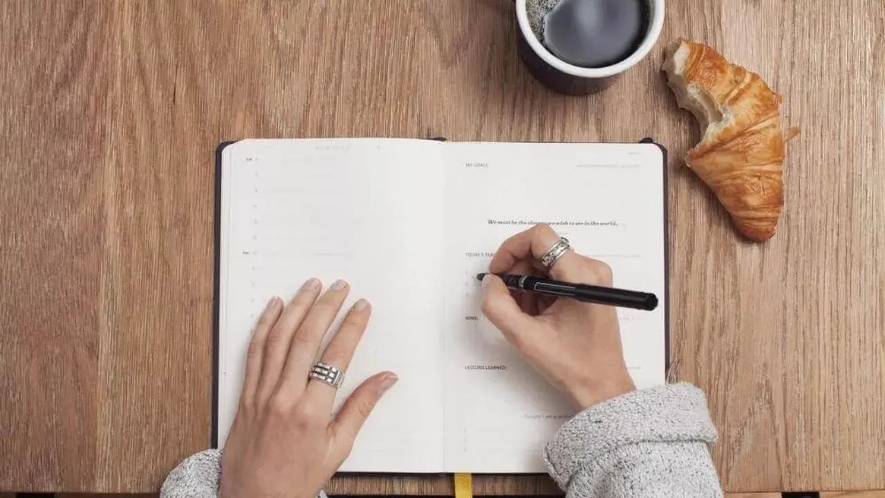 A person's hand holding a pen, writing in a notebook that is open on a wooden table