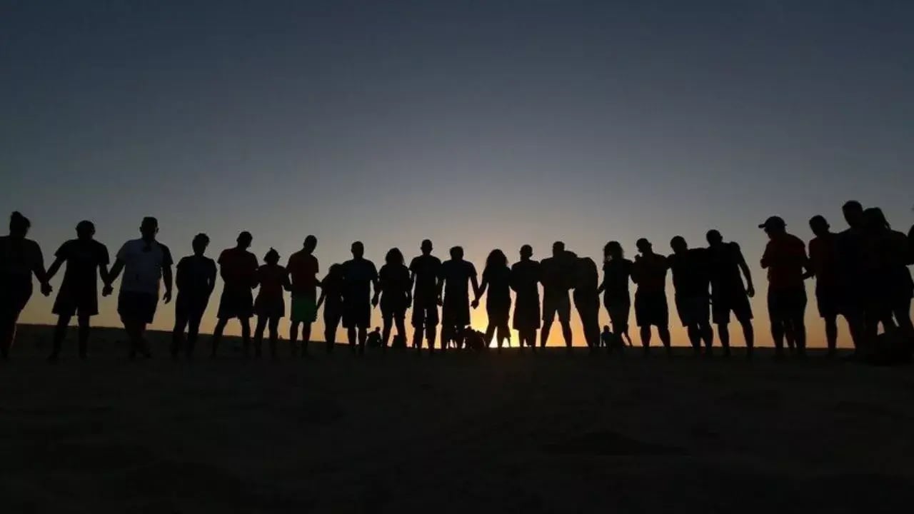 The image depicts a group of people standing in a line, holding hands and forming a silhouette against a vibrant sunset in a desert setting.