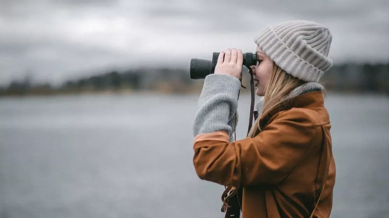 A person wearing a beanie and coat is looking through binoculars over a body of water.