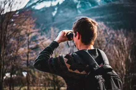 The image shows a person from behind, wearing a black jacket and a backpack, taking a photograph with a camera.