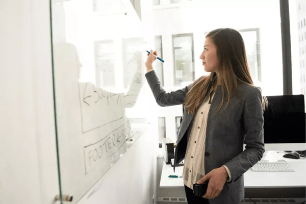 A person in a business casual outfit is writing on a whiteboard in an office environment.