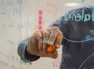 The image shows a person's hand holding a marker, writing on a whiteboard that has various words and equations written in different colors.