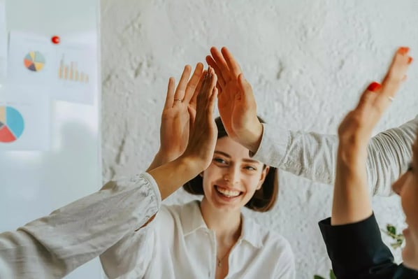Three people are standing in front of a white wall with a poster of charts and graphs. They are clapping their hands together in a synchronized manner.