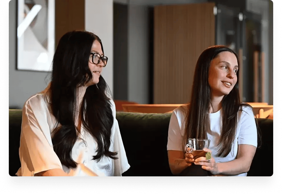 Two women are sitting on a couch, one holding a glass with a beverage, and both are dressed in casual attire.